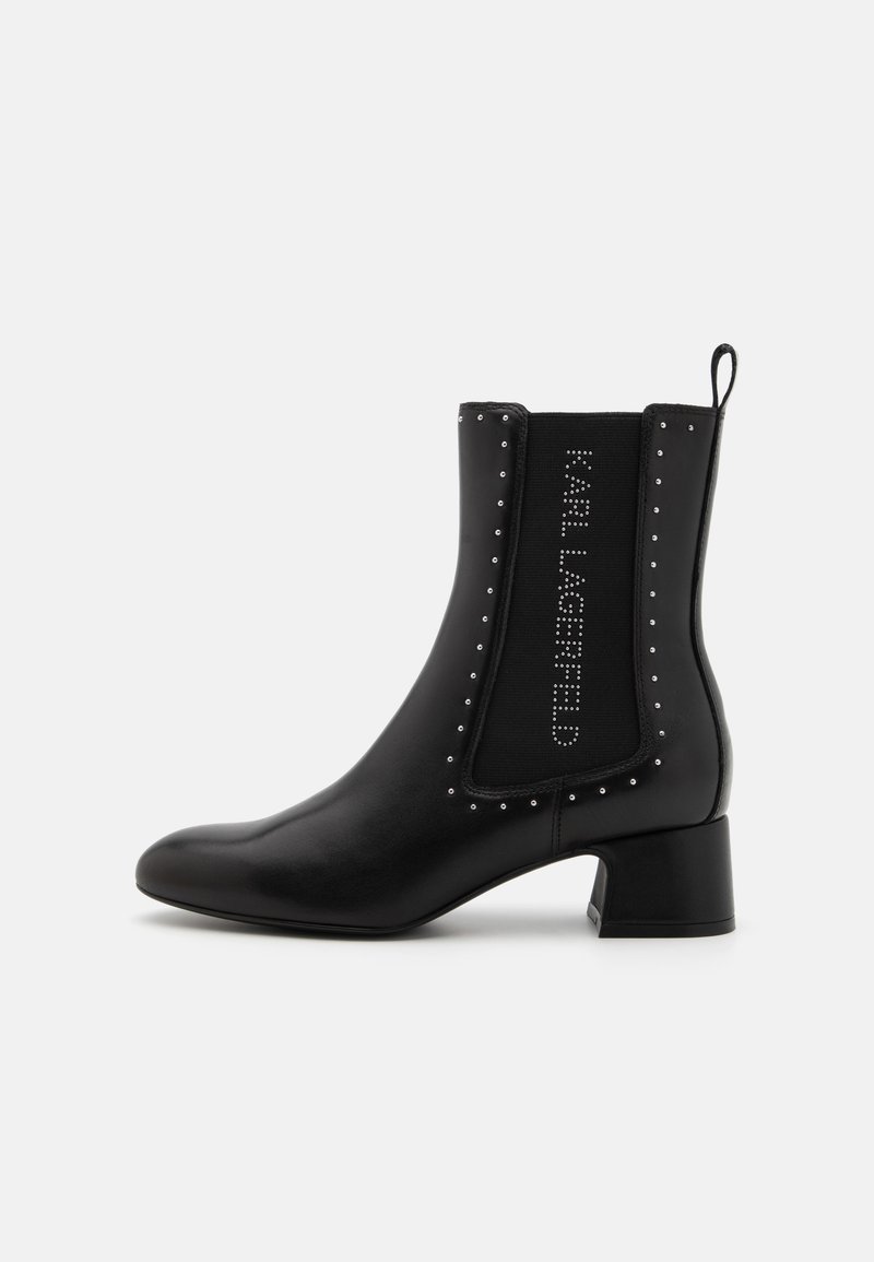 Botín negro hasta el tobillo con punta afilada, hecho de cuero suave. Presenta un panel elástico y texturizado con tachuelas y un pequeño tacón cuadrado.