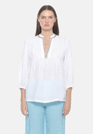 Donna con capelli castani lisci indossa una camicetta bianca a maniche lunghe con pieghe e collo decorato con perline, abbinata a pantaloni azzurri, di fronte.