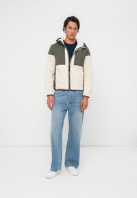 Fleecejakke med grønn og kremfarge blokkering, glidelås foran, to-farget design, brukt over en marineblå skjorte, kombinert med lyseblå jeans.