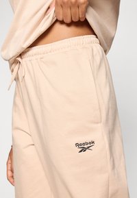 Gaiši persiku krāsas Reebok sporta bikses ar elastīgu jostu, auklu, sānu kabatām un melnu izšūtu logotipa detaļu. Mīksts, viegls audums.