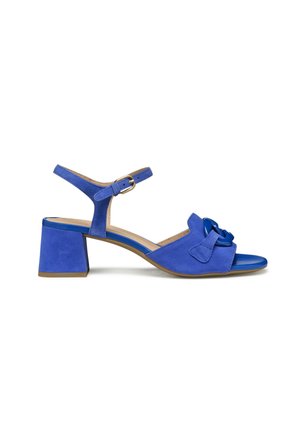 Sandal azul de ante con tacón cuadrado, punta abierta y correa ajustable en el tobillo; cuenta con un detalle de lazo en la parte delantera. Suela plana y bordes redondeados.