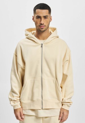 Beige zip-up hoodie in soft fabric with a large hood, kangaroo pockets, and relaxed fit. Features minimalist design and ribbed cuffs.