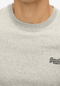 Gros plan sur le cou et le haut de la poitrine d'une personne portant un t-shirt rayé gris clair avec un petit logo "SuperDry" sur le côté droit.