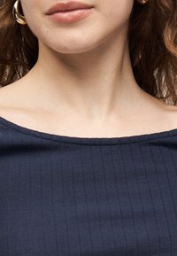 Γυναίκα που φοράει χρυσούς κρίκους σκουλαρίκια και μπλε ναυτικό κορυφή με ριπ υφή και ακάλυπτους ώμους, που δείχνει τον λαιμό και το πάνω μέρος του στήθους.