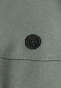 Detailní záběr šedo-zelené látky s prošitým švem a kulatou černou pryžovou nášivkou s abstraktním geometrickým vzorem.