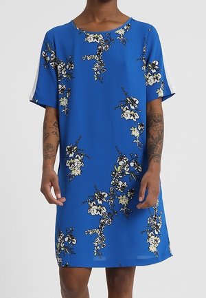 Blaues Kurzarmkleid mit weißem und schwarzem Blumenmuster, getragen von einer tätowierten Person vom Hals bis zu den Knien.
