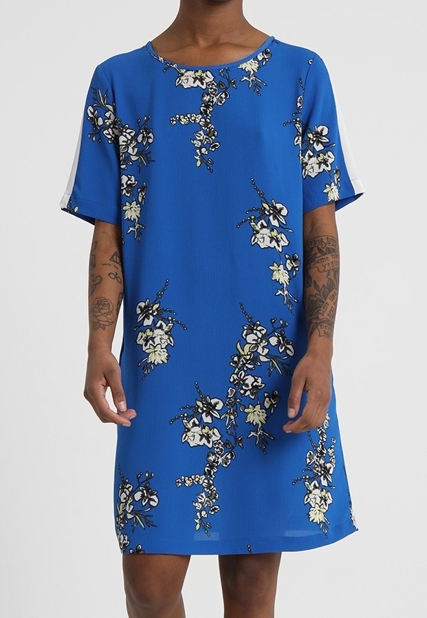 Robe bleue à manches courtes avec imprimé floral blanc et noir portée par une personne tatouée du cou aux genoux.