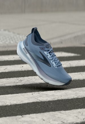 Světle modrá běžecká bota Brooks s tlustou bílou podrážkou, která visí nad černobílým přechodem na asfaltu.