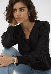 Chemisier noir à manches longues avec des manches bouffantes et un décolleté en V. Le tissu a une finition texturée. Associé à un jean bleu et un bracelet argenté brillant.