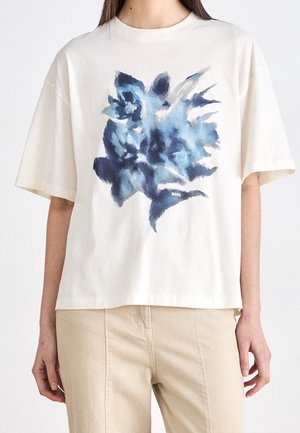 Biała koszulka z krótkim rękawem z abstrakcyjnym niebieskim kwiatowym nadrukiem i małym logo "BOSS", noszona przez stojącą osobę z beżowymi spodniami.