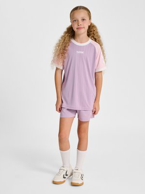 Junges Mädchen mit langen lockigen Haaren, das rosa und lilafarbene Sportkleidung, weiße Socken und weiße Sportschuhe trägt, steht vor einem schlichten Hintergrund.