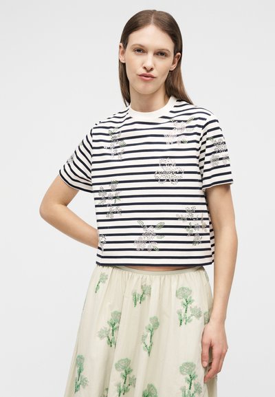 Γυναίκα που φοράει λευκό και ναυτικό μπλε ριγέ κοντομάνικο τοπ με λουλουδάτα μοτίβα, συνδυασμένο με κρέμ φούστα με πράσινη λουλουδένια κεντητική.