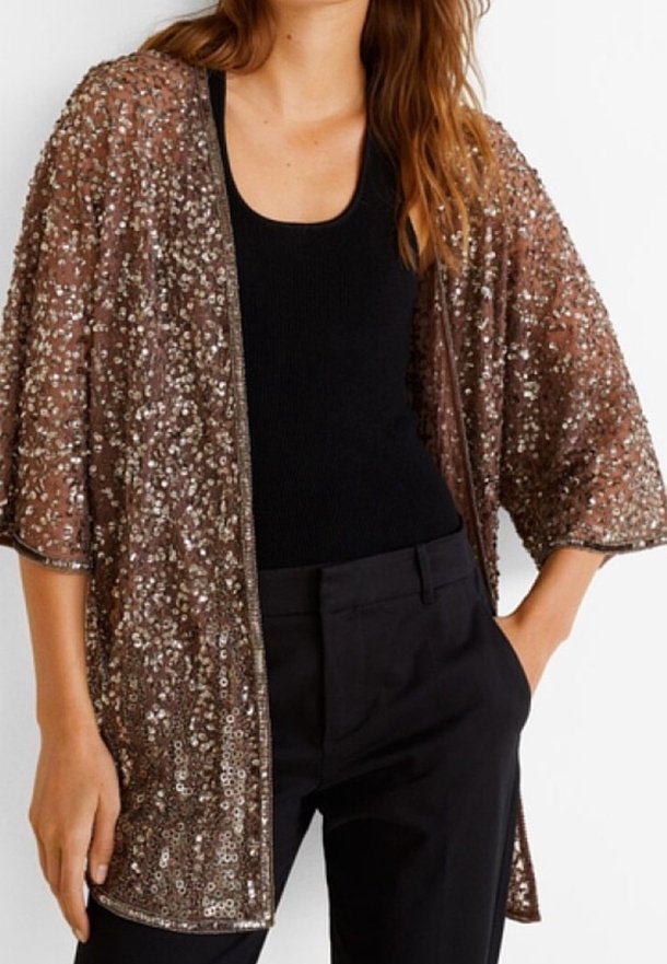 Femme portant une veste marron à sequins ouverte sur un top noir sans manches et un pantalon noir, une main dans la poche.