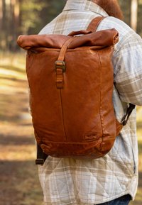 Sac à dos en cuir marron à rabat roulant avec des bretelles réglables et une fermeture à boucle. Présente une texture lisse et un design minimaliste avec un détail de logo.
