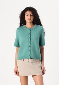 Short-sleeve, teal cardigan with a round neck, featuring a button-up front and ribbed hem and cuff detailing. Worn over a beige skirt.