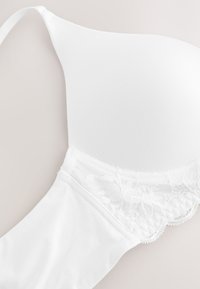 Weißer BH mit glatten, geformten Cups und Spitzendetail nahe dem Unterbrustbereich vor neutralem Hintergrund.