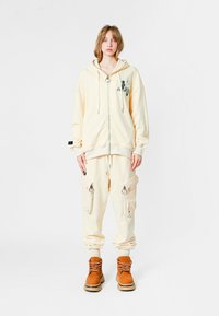 Una persona joven está de pie vistiendo una sudadera oversized de color beige claro y pantalones cargo con grandes bolsillos, combinados con botas de cordones color marrón claro, sobre un fondo blanco.