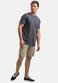 T-shirt en coton gris à encolure ronde, associé à un short beige. Le modèle porte des chaussures en toile noires avec des lacets blancs, se tenant dans une pose neutre.