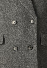 Dvřevý šedý vlněný kabát s měkkou texturou, s velkými stříbrnými knoflíky a dvěma bočními kapsami. Čisté linie a klasický design.