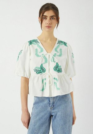 Blusa blanca de tela ligera, con bordado floral verde, mangas cortas abullonadas, cuello en V y cintura ajustable con cordón.