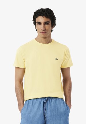Giovane uomo con capelli ricci scuri che indossa una maglietta giallo chiaro a maniche corte e pantaloni blu con coulisse, in piedi con le mani nelle tasche.