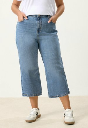 Persoon draagt lichtblauwe cropped wide-leg jeans, witte sneakers met zwarte strepen en een wit shirt, met de handen in de zakken, staand op een lichte vloer.