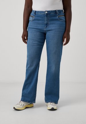 Jeans en denim bleu à taille mi-haute et évasée, avec des poches avant et une texture subtile. Portés avec des chaussures de sport de couleur claire.
