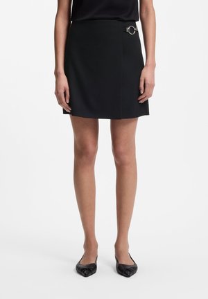 Vrouw draagt een zwarte minirok met een zilveren gespdetail in de taille, gecombineerd met zwarte platte schoenen, staand tegen een effen achtergrond.
