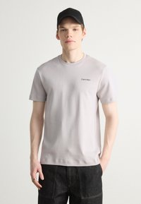 Camiseta de algodón gris claro con cuello redondo, mangas cortas y un pequeño logotipo negro de "Calvin Klein" en el lado izquierdo del pecho.