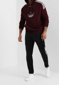 Man som bär en vinröd Adidas-hoodie med vita ränder och logotyp, svarta Adidas-träningsbyxor och vita sneakers som går inomhus.