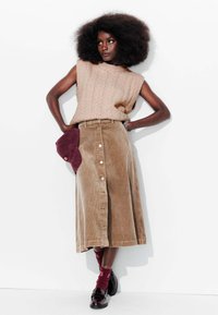 Pull en maille beige sans manches, jupe boutonnée en velours côtelé marron, chaussettes bordeaux, chaussures noires et casquette bordeaux. Style simple et décontracté.