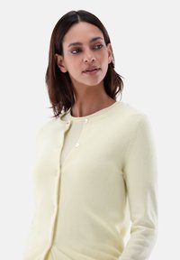 Helles gelbes Strickjacke aus weichem Material, mit einem runden Ausschnitt, langen Ärmeln und weißen Knöpfen entlang der Vorderseite.