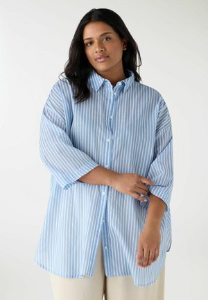 Donna con pelle media e capelli scuri che indossa una camicia a righe verticali larghe azzurro chiaro e bianco con bottoni e maniche lunghe, abbinata a pantaloni chiari.