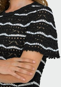 Sort-hvid stribet hækletrøje med bølgende mønstre, rund halsudskæring og scallop-korte ærmer. Struktureret åbent strikdesign.
