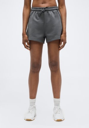 Persona con pantalones cortos de cordón gris oscuro, calcetines blancos y zapatillas deportivas blancas, de pie frente a un fondo claro y sencillo.