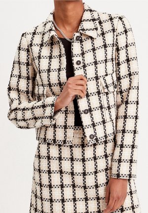 Personne portant une veste texturée à carreaux beige et noire et une jupe assortie, avec un haut noir et un collier chaîne en argent.