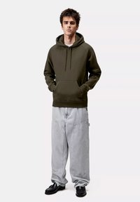 Felpa verde oliva con tasca a marsupio, abbinata a pantaloni grigi chiari oversize con tasche laterali e completata da scarpe nere.