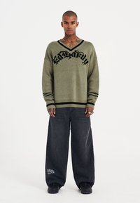 Maglione verde a maglia con lettering nero, maniche larghe e dettagli a strisce nere. Abbinato a jeans neri larghi e sneakers scure.