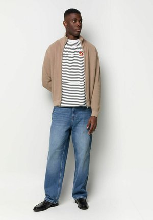 Mann steht vor einer weißen Wand und trägt einen beigen Zip-Up-Pullover, ein gestreiftes weiß-schwarzes T-Shirt, lockere blaue Jeans und schwarze Schuhe.