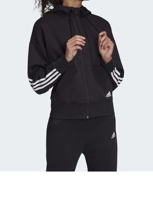 Osoba ubrana w czarny dres Adidas z białymi, trzema paskami na rękawach i logo Adidas, regulująca sznurki kaptura.