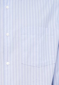 Ανοιχτό μπλε πουκάμισο με κάθετες λευκές ρίγες, με τσέπη στο στήθος και ομαλή υφή υφάσματος. Τα κουμπιά είναι λευκά και στρογγυλά.