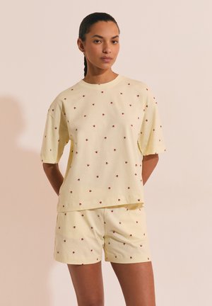 Femme aux cheveux tressés portant un ensemble chemise et short assorti jaune clair avec de petits imprimés de tranches de pastèque, debout devant un fond uni.