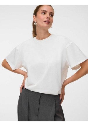 Femme portant un t-shirt blanc ample à manches courtes rentré dans un pantalon plissé gris foncé taille haute avec des boucles d'oreilles anneaux.