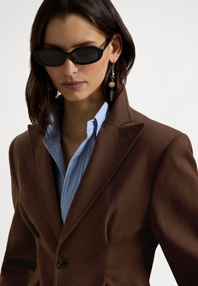 Καφέ προσαρμοσμένο σακάκι με ένα κουμπί, συνδυασμένο με ανοιχτό μπλε πουκάμισο με ρίγες. Μαύρα γυαλιά ηλίου και κρεμαστά σκουλαρίκια ολοκληρώνουν την εμφάνιση.