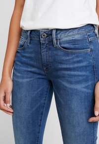 Blå denimjeans med smal passform, femfickorsdesign och knappstängning. Mjuk textur med lätt slitage och sömnadsdetaljer.