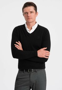 Schwarzer V-Ausschnitt-Pullover aus weichem Material, kombiniert mit einem weißen Hemd mit Kragen. Graue Hose mit einem schwarzen Gürtel, der ein strukturiertes Gewebe aufweist.