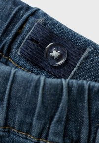 Tessuto di denim blu con cintura elastica, dotato di un piccolo bottone trasparente e dettagli di cucitura. Primo piano dell'area della cintura.