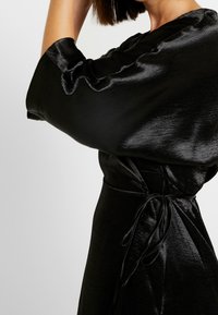 Mustasilkkinen mekko, jossa on leveät hihat ja solmittava vyötäröyksityiskohta. Kangas on kiiltävä ja sileä, korostaen eleganttia siluettia.