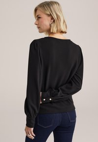 Rövid szőke hajú nő, fekete, hosszú ujjú, texturált blúzt és sötétkék farmert visel, egyik kezét a háta mögött tartva áll.