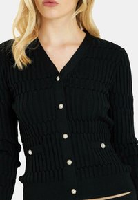 Schwarze gerippte Strickjacke mit V-Ausschnitt, Perlmuttknopf-Verschlüssen und zwei Vordertaschen mit geripptem Saum. Strukturierter Strickmuster über die gesamte Jacke.